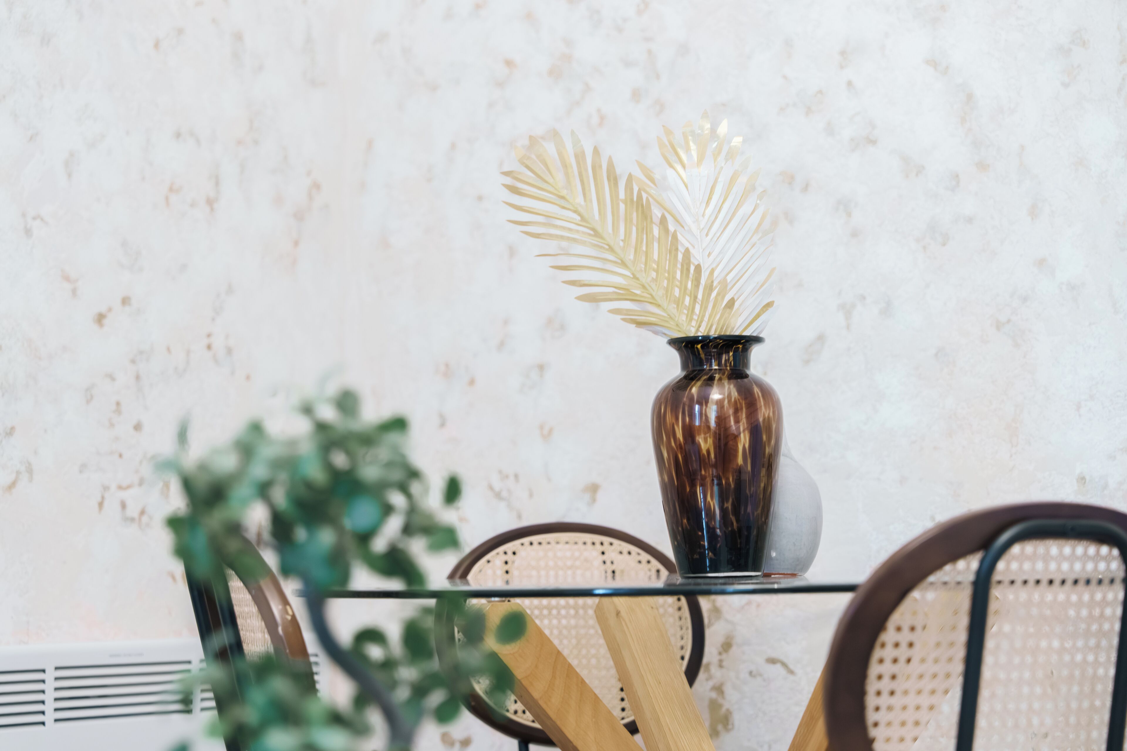 A dining table with a glass top, surrounded by wicker chairs, and a decorative vase with a plant.