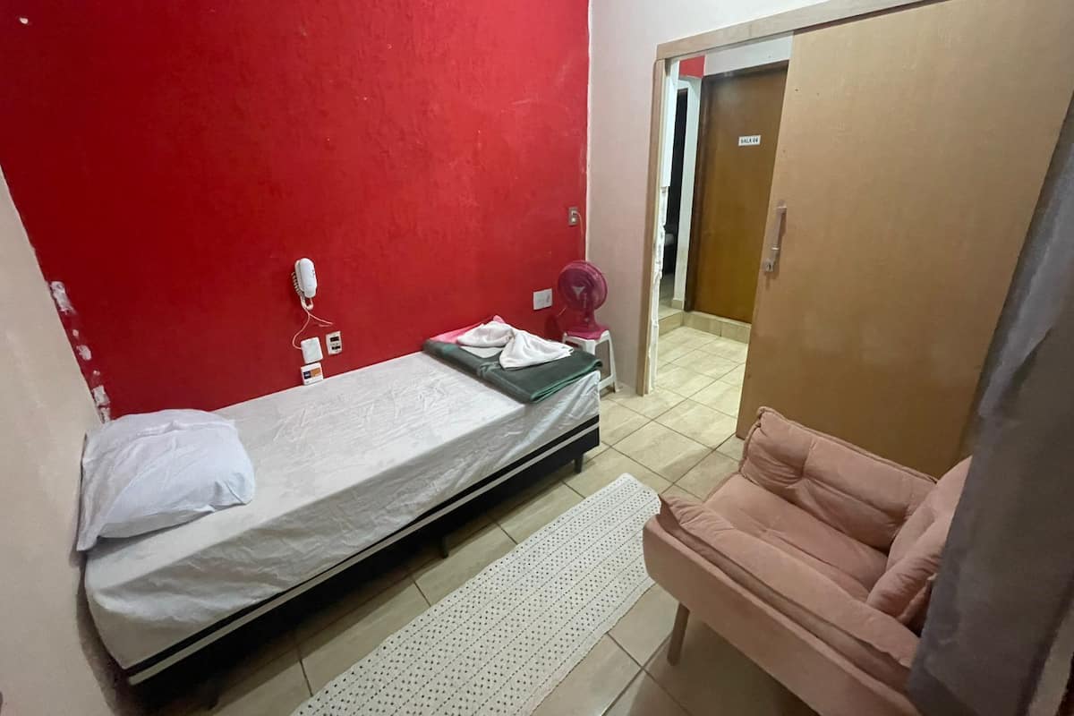 A small, single-bed room with a red wall, a wooden door, a pink chair, and a white bedside table.