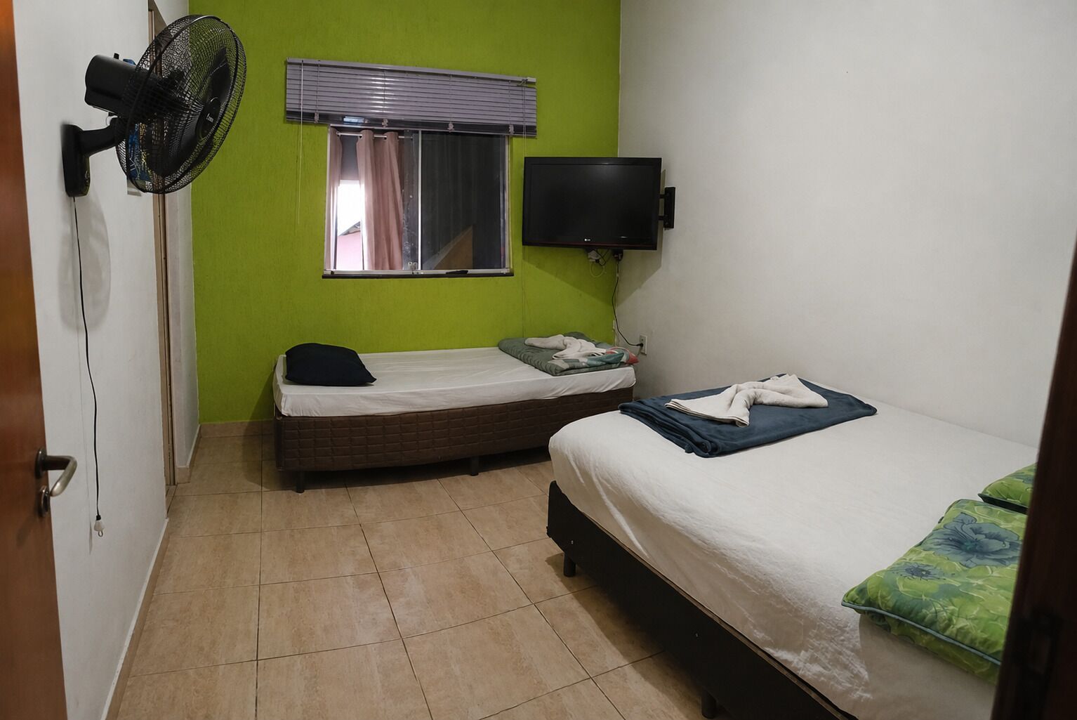 Une petite chambre d’hôtel composée d’une seule pièce, comprenant un lit, un téléviseur, une fenêtre avec des stores, un ventilateur et une porte.