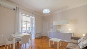 A modern living room with a wooden herringbone floor, a white dining table with chairs, a sofa with floral cushions, a small white table, and a kitchen area with a white cabinet and a hanging light fixture. - Sea View Central Apartment by Wonderful Italy (Sanremo)