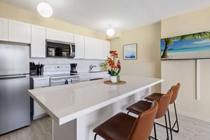 A modern kitchen with white cabinetry, a stainless steel refrigerator, a microwave, and a coffee maker. There is a small island with a vase of flowers and two bar stools. - 20th Fl Waikiki Banyan | King | AC | Parking (Honolulu)