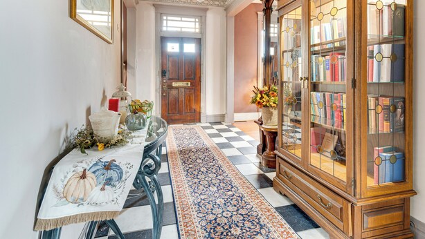 Um corredor com piso xadrez, uma mesa com toalha, um armário de madeira com portas de vidro e uma porta ao fundo.