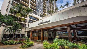 A hotel entrance with a covered walkway, palm trees, and a view of a high-rise building in the background. - 29th Fl Waikiki Banyan | Diamond Head Views | Parking | AC (Honolulu)