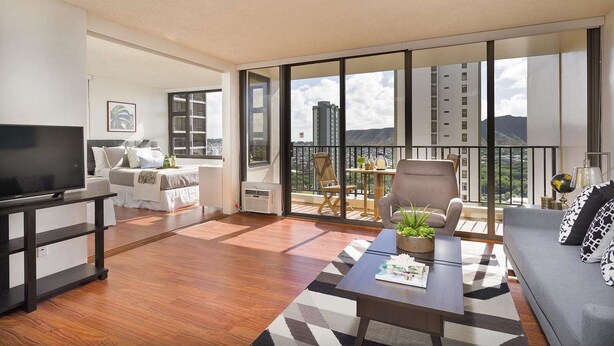 A modern living room with a flat-screen TV, a sofa, a coffee table with a magazine, and a balcony with a view of the city and mountains. - 29th Fl Waikiki Banyan | Diamond Head Views | Parking | AC (Honolulu)