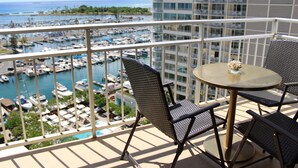 Ein Balkon mit Blick auf einen Yachthafen und den Ozean. Zwei Stühle und ein Tisch mit einer Vase sind vorhanden.