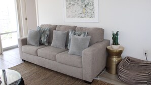 Ein Wohnzimmer mit einem beigefarbenen Sofa, gemusterten Kissen, einem gerahmten Kunstwerk, einer Topfpflanze und einem gewebten Pouf.