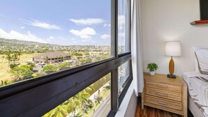 Une chambre d’hôtel dotée d’une grande fenêtre donnant sur un parking et des immeubles. On y trouve une table de chevet avec une lampe et une petite plante.
