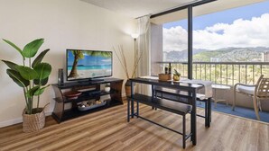 Un salon moderne doté d’un téléviseur, de parquet, d’un balcon offrant une vue sur la ville et les montagnes, ainsi que d’un coin repas avec une table et des chaises.