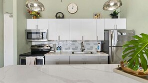 A modern kitchen with white cabinets, stainless steel appliances, a microwave, oven, and refrigerator. There is a sink with a faucet, a clock on the wall, and a potted plant on the counter. - 38th Fl Waikiki Banyan | King | AC | Parking (Honolulu)