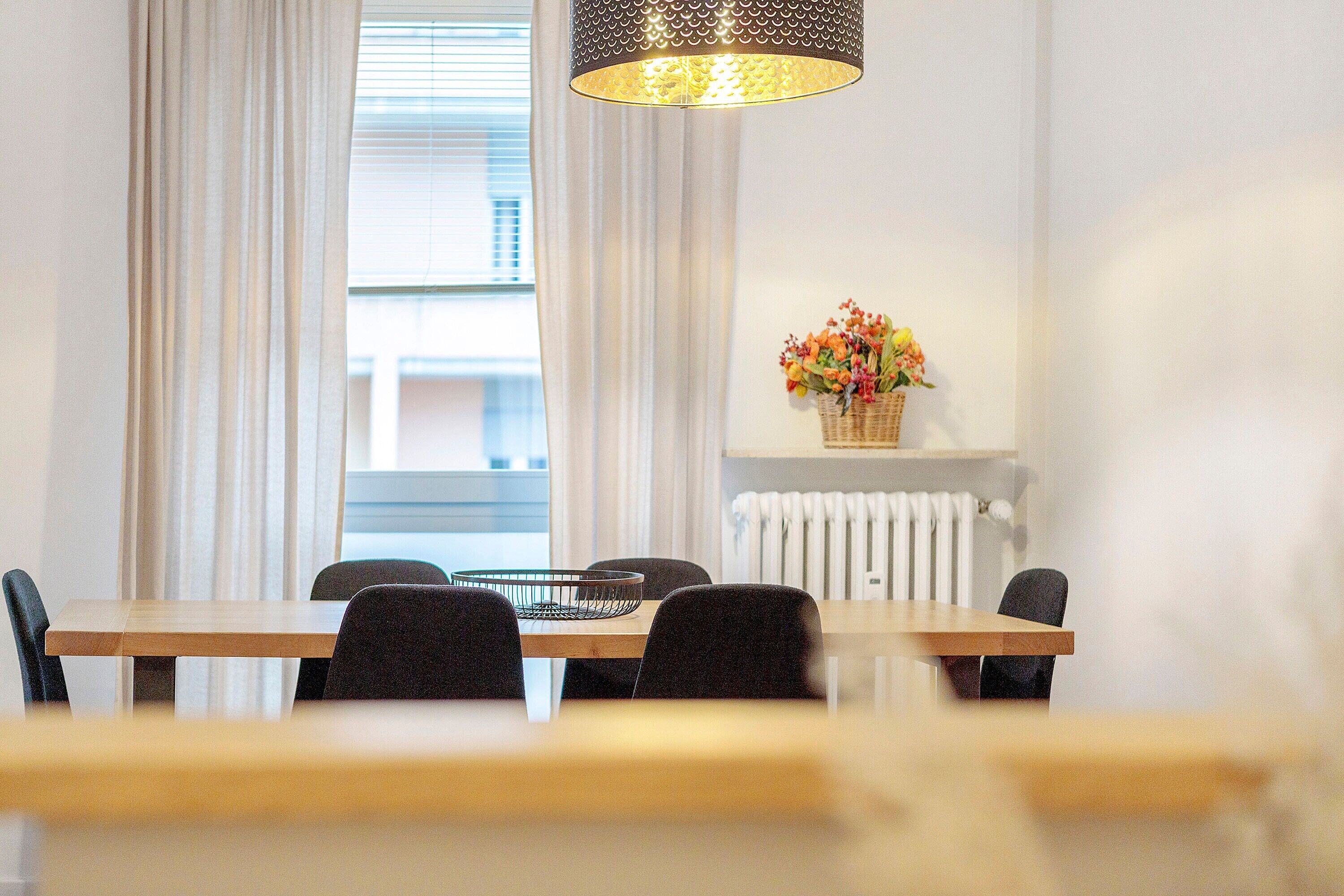 A dining area with a table, chairs, a basket of flowers, and a window with curtains.