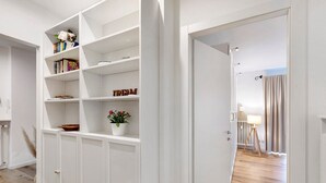 Un intérieur aux murs blancs, avec une bibliothèque, une plante en pot et une porte donnant sur une autre pièce.