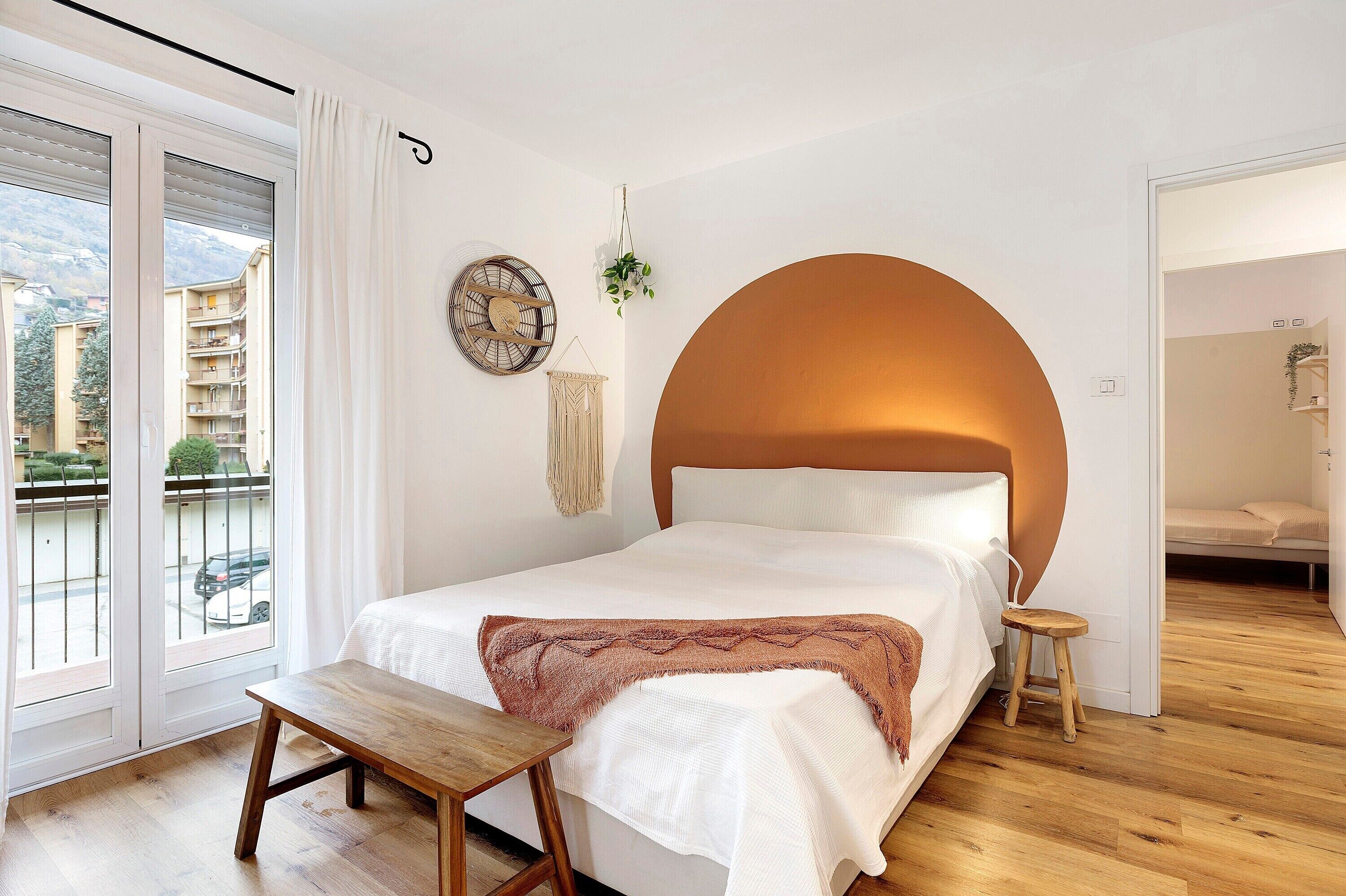A bedroom with a bed, a wooden bench, a large window with a view, and a circular wall design.