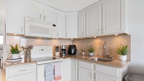 A modern kitchen with white cabinetry, a built-in microwave, a coffee maker, and a sink. There are potted plants on the counter. - 7th Fl Aloha Lani  | Lanai | AC | Parking | Pool (Honolulu)