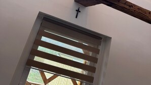 A room with a sloped ceiling, exposed wooden beams, a window with a view, a wall-mounted clock, and a door with horizontal blinds. - The Hungarian Valley (Szellő)