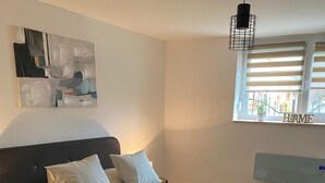 A modern bedroom with a large bed, a headboard, a wall-mounted heater, a window with blinds, and a hanging light fixture. - The Hungarian Valley (Szellő)