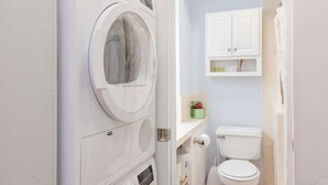 Une petite salle de bain équipée d’un lave-linge à chargement frontal, de toilettes et d’un petit meuble contenant des articles de toilette.