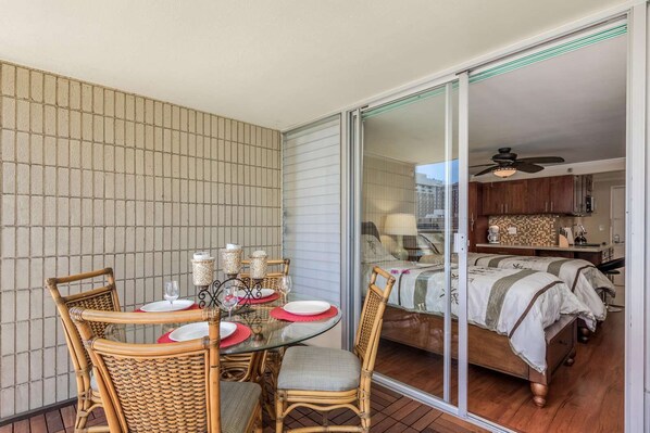 A balcony with a glass table set for dining, wicker chairs, and a view into a bedroom with a bed and a small kitchenette. - 9th Fl Island Colony Studio | Balcony | Pool (Honolulu)