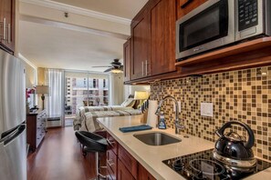 A modern kitchen with a microwave, stove, and sink. There is a dining area with a table and chairs, and a view of a balcony with a railing. - 9th Fl Island Colony Studio | Balcony | Pool (Honolulu)