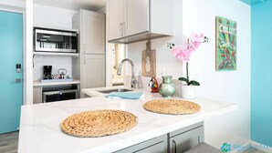 Una cuina moderna amb armaris blancs, una campana extractora d'acer inoxidable i una cafetera. Hi ha dos roba de taula teixits al taulell, un vas amb flors roses i un senyal decoratiu amb forma de pinya.