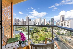 A balcony with a view of a cityscape, a wicker chair, a small table with a tray of flowers, and a decorative wall panel. - 15th Fl Luana Waikiki Studio | City & Mountain Views | Pool | Balcony (Honolulu)