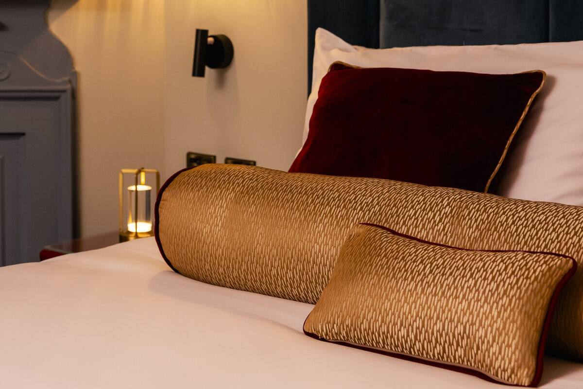 A neatly made bed with a golden woven headboard and pillows, a red accent pillow, and a small table with a lamp beside it.