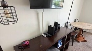 Un salon doté d’un téléviseur à écran plat fixé au mur, d’un meuble de rangement en bois foncé placé en dessous, d’une petite table avec une lampe et d’un sol recouvert d’une moquette.