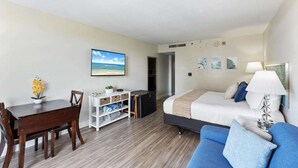 A hotel room with a bed, a blue sofa, a dining table with chairs, a small desk, and a painting of a beach scene. - 3rd Fl Palms Waikiki Studio Overlooking the Pool | Parking (Honolulu)