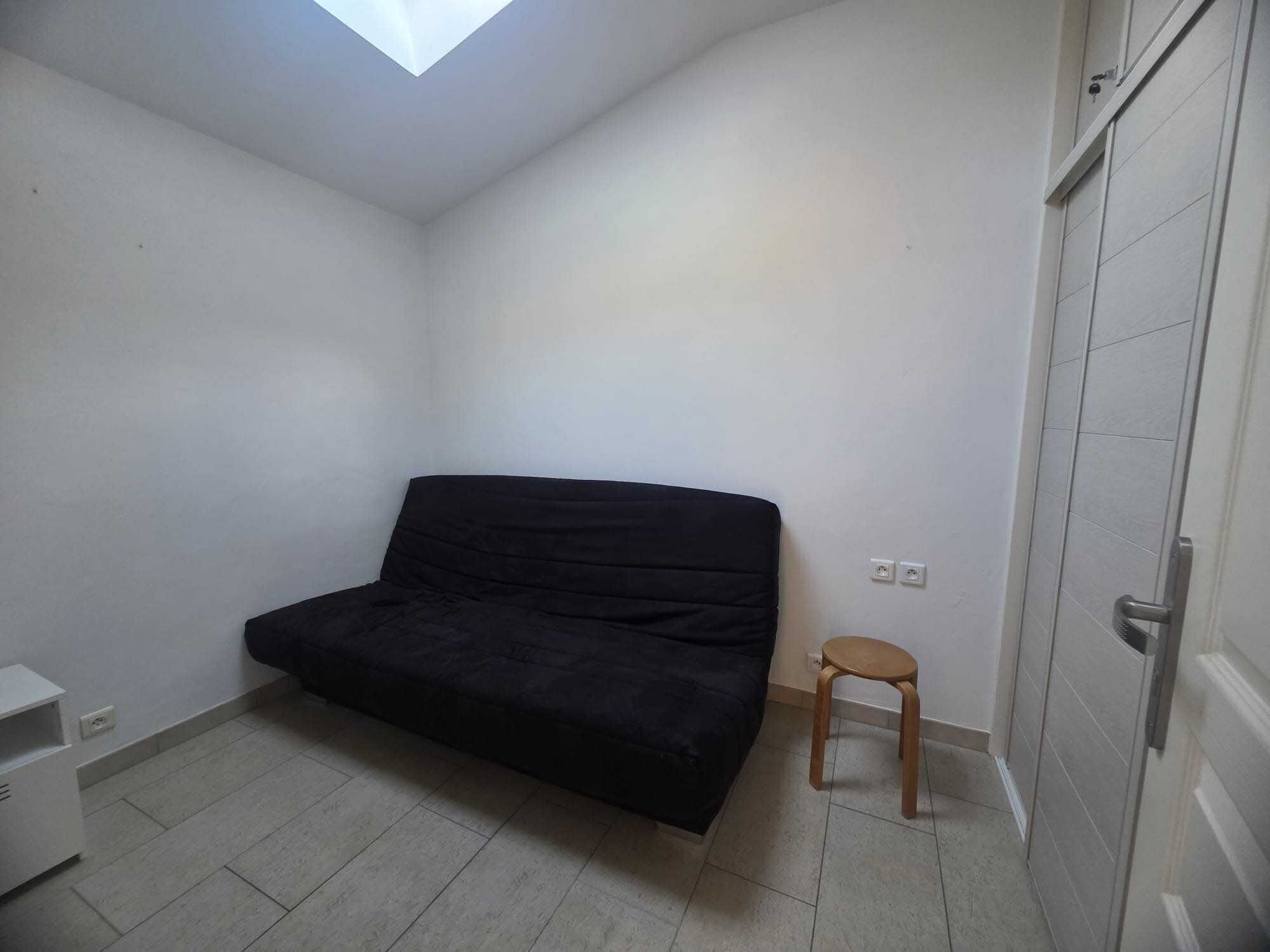 Una habitación pequeña y escasamente amoblada con un sofá negro, un taburete de madera y un armario blanco.