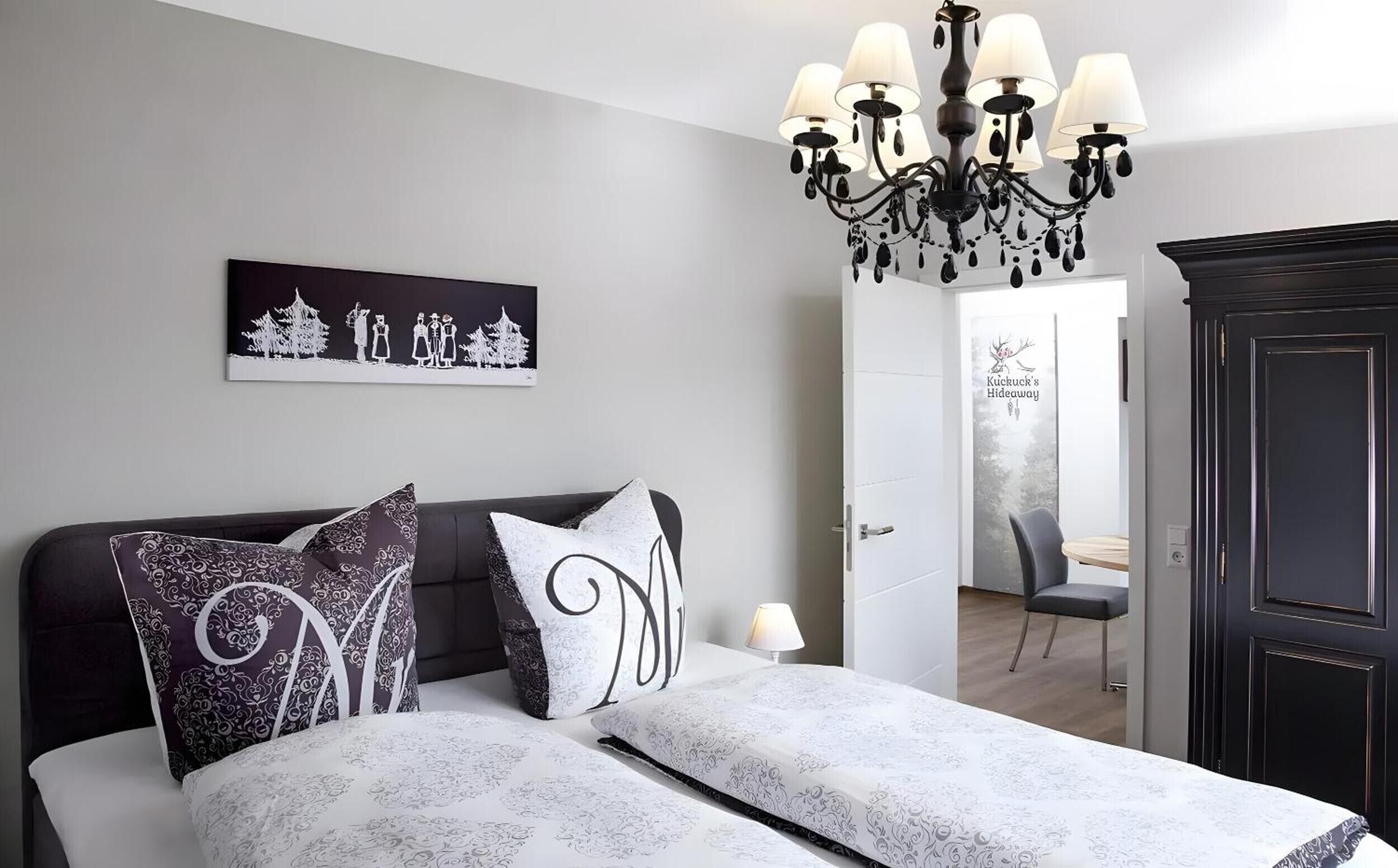 A bedroom with a bed, a chandelier, a painting, a wardrobe, and a door leading to another room.