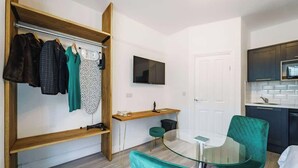 Un espace de vie compact comprenant un coin cuisine, un espace repas avec une table en verre et des chaises turquoise, ainsi qu’un portant à vêtements avec divers articles.