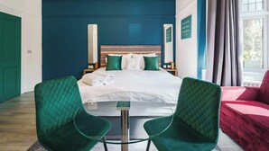 Une chambre d’hôtel avec un lit, deux fauteuils verts, une petite table et un canapé rouge.