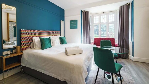 Une chambre d’hôtel avec un grand lit, un bureau avec un miroir, une petite table et une chaise. Il y a une fenêtre avec des rideaux et une vue sur l’extérieur.