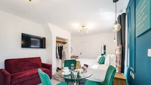 Moderni hotellihuone, jossa on punainen sohva, turkoosinsiniset tuolit, lasipöytä ja valkoisilla lakanoiden päällisillä varustettu sänky.