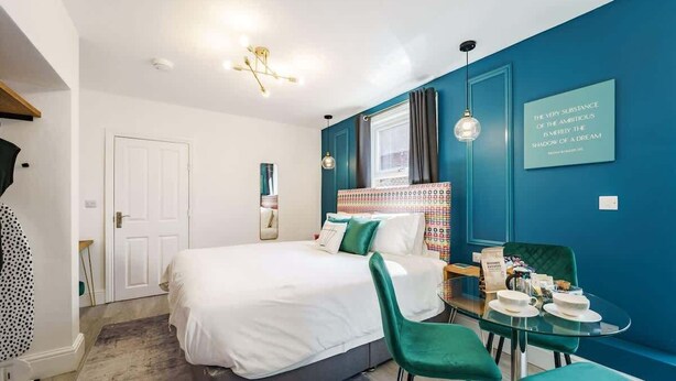 Moderni hotellihuone, jossa on sänky, lasinen pöytä tuoleineen, sininen tehosteseinä ja inspiroiva sitaatti.