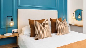 Una cama con ropa de cama blanca y almohadas a rayas, una mesita de noche con lámpara y una pared con diseño de panel azul.