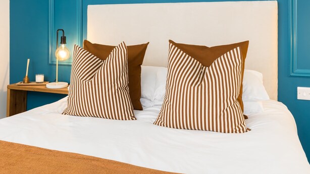 Una cama bien tendida, con almohadas a rayas y una mesita de noche con una lámpara y una vela.