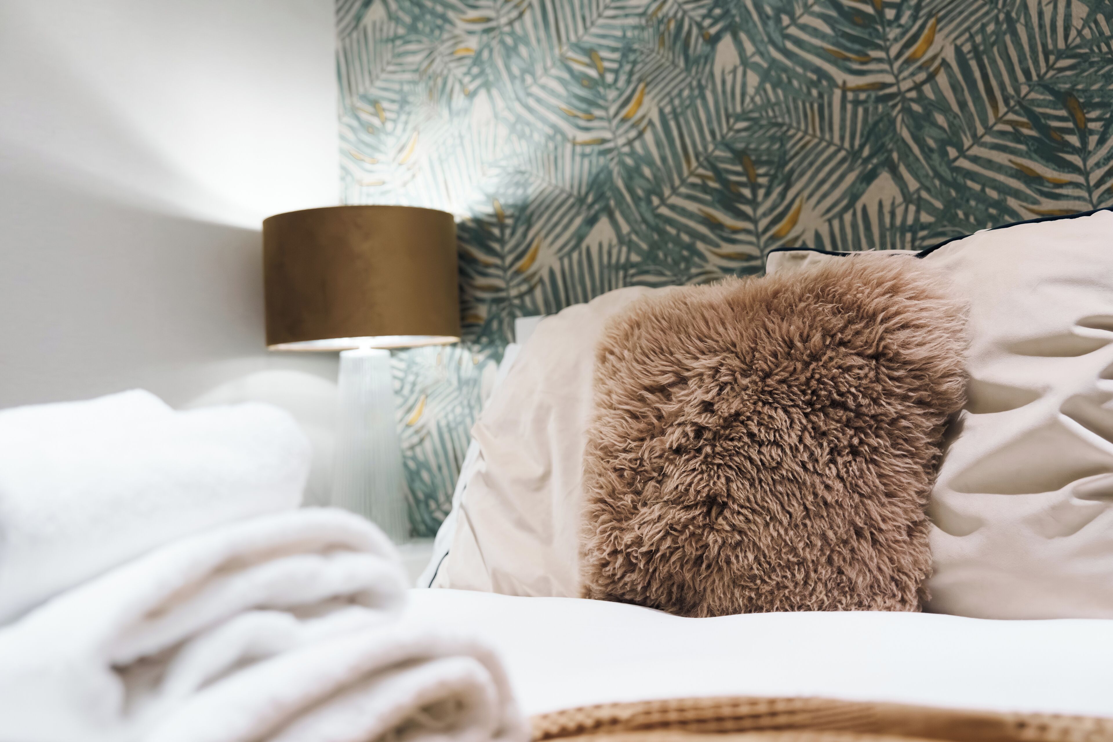 A neatly made bed with a fluffy pillow, a lamp, and a wall with a leafy pattern.