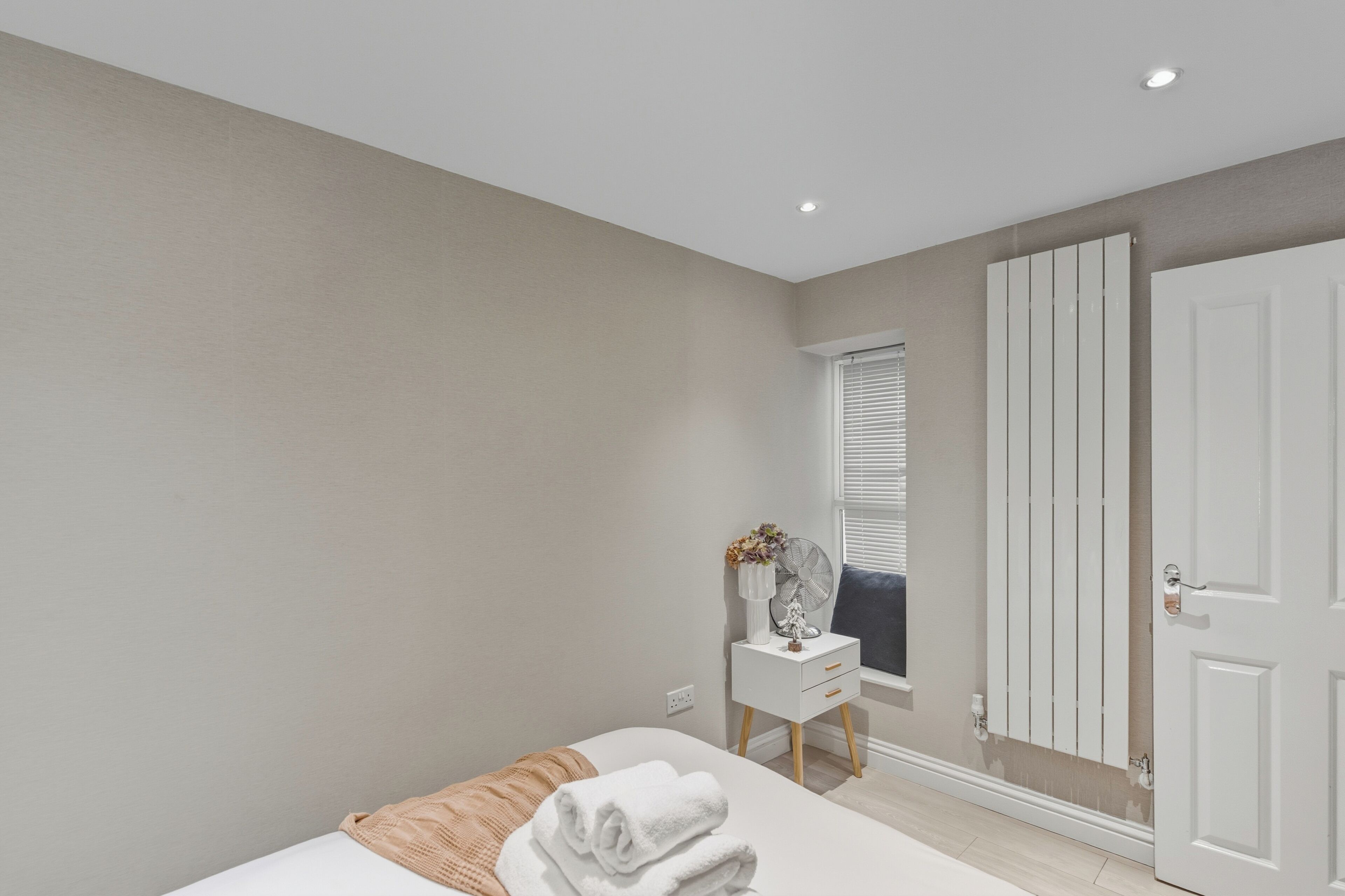 Un dormitorio con cama, mesita de noche con ventilador, ventana con persianas y una puerta blanca.