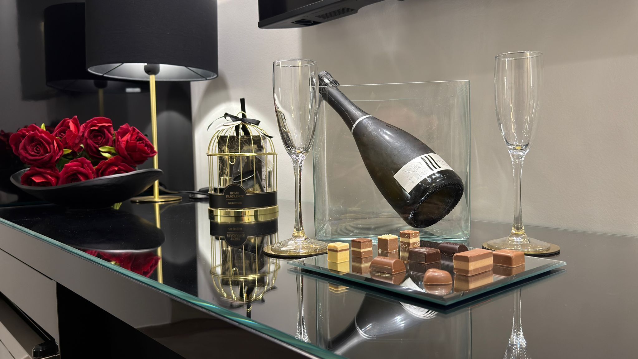 Une table à plateau de verre sur laquelle se trouvent une bouteille de champagne, un verre, un flacon de parfum et des chocolats.