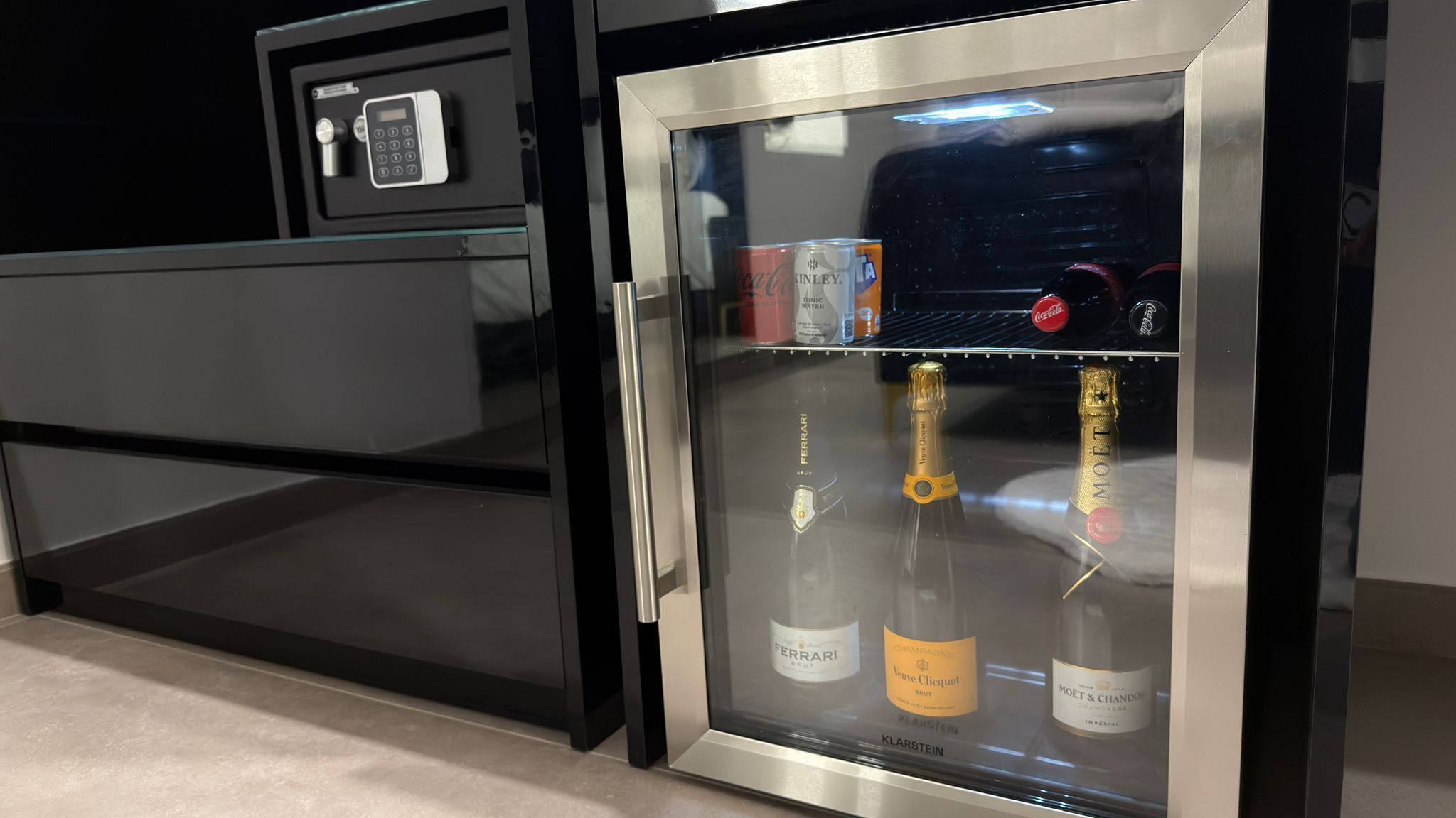 Un minibár con puerta de vidrio, que contiene una botella de champán, una botella de vino y una lata de Coca-Cola.