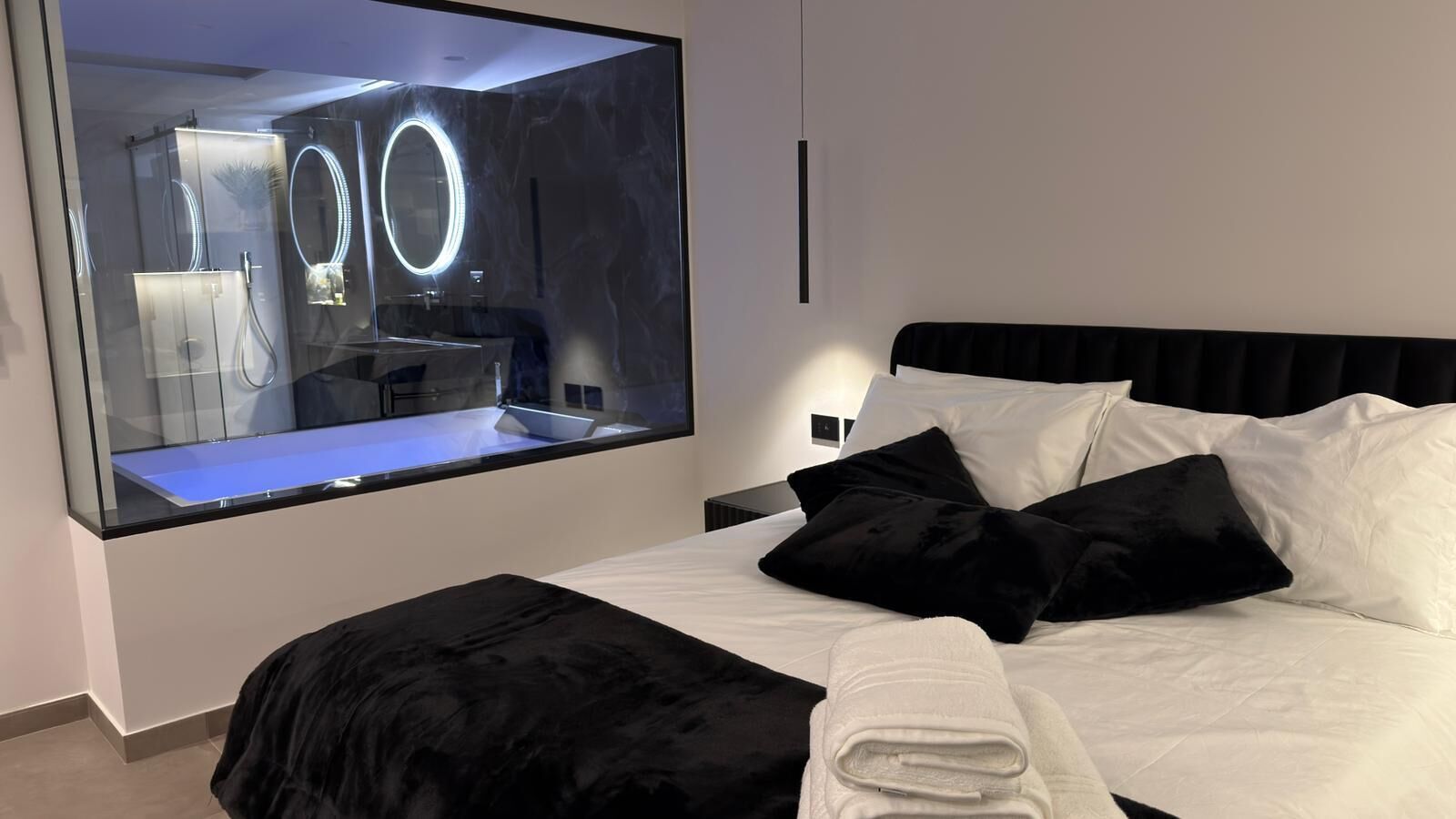 Une chambre d’hôtel moderne avec un grand lit, des oreillers noirs et blancs, un pouf noir et un grand miroir doté d’anneaux lumineux.