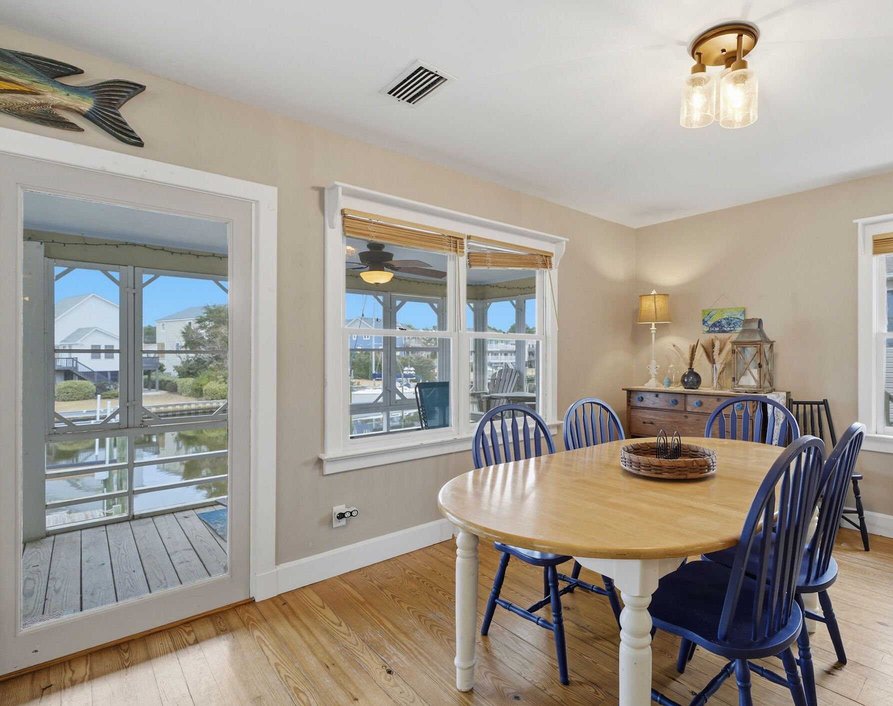 A dining area with a wooden table and blue chairs, large windows with a view of a dock and boats, and a ceiling fan.