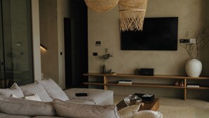A modern living room with a large sectional sofa, two woven pendant lights, a flat-screen TV, and a minimalist wooden coffee table. - Lithos villa by Elements villas (Kokkino Chorio)