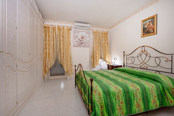 Un dormitorio con cama, armario, cortinas, un cuadro y una mesita de noche.