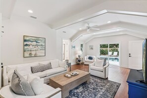 Ein geräumiges Wohnzimmer mit einem weißen Sofa, einem Holztisch und einem blau gemusterten Teppich. Es gibt einen Deckenventilator, ein Bild an der Wand und einen Blick auf einen Pool durch große Fenster.