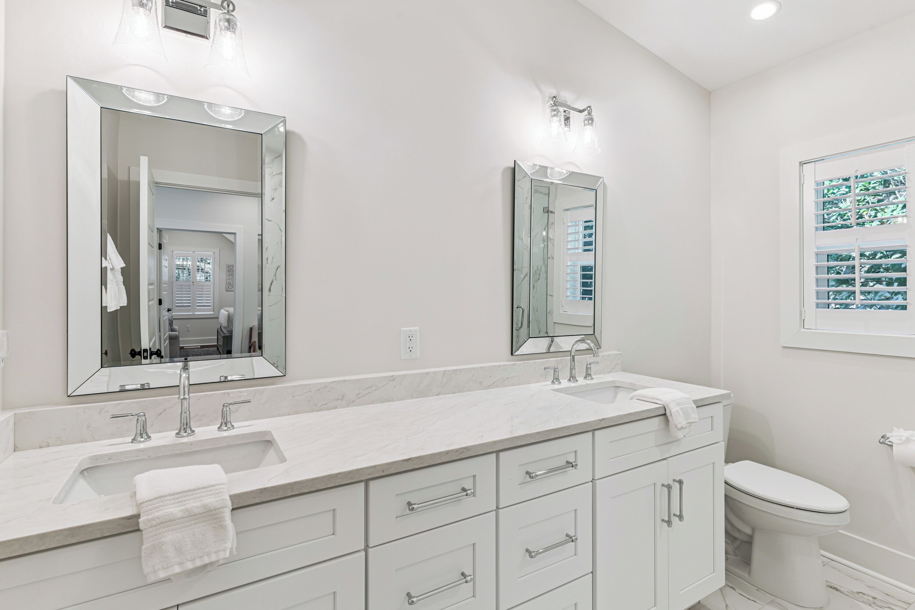 Une salle de bain moderne avec un grand miroir, deux lavabos, des toilettes et une fenêtre équipée de stores.