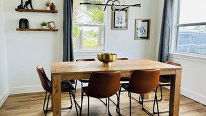 Et spiseområde med et træbord, fire stole og en skål i messing. To hylder med dekorative genstande og et vindue med gardiner.