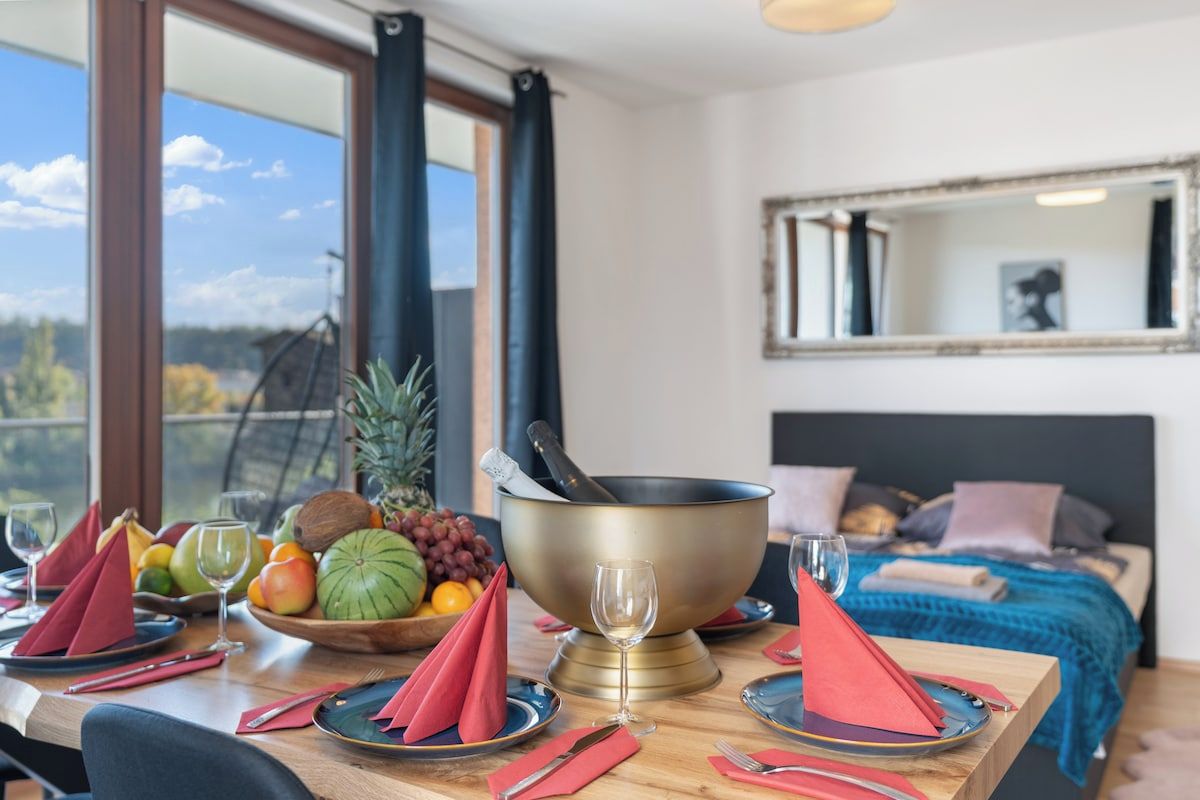 Een eettafel gedekt met rode servetten, borden en een schaal fruit. In de achtergrond is een bed met een blauwe deken en kussens te zien.