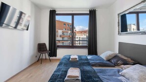 Ett modernt hotellrum med ett stort fönster, en platt-TV, en säng med ett blått överkast och en stol.