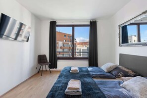 A modern hotel room with a large window, a flat-screen TV, a bed with a blue bedspread, and a chair. - Terrace ONE (Prague 7)